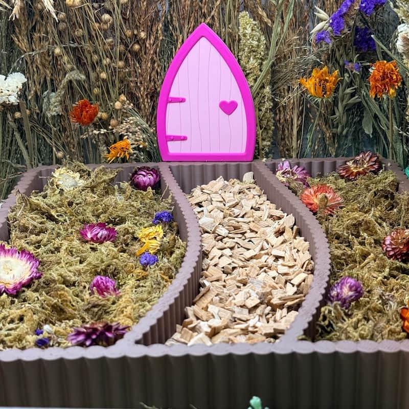 Pink fairy door on a hamster garden with flowers and wood chips.